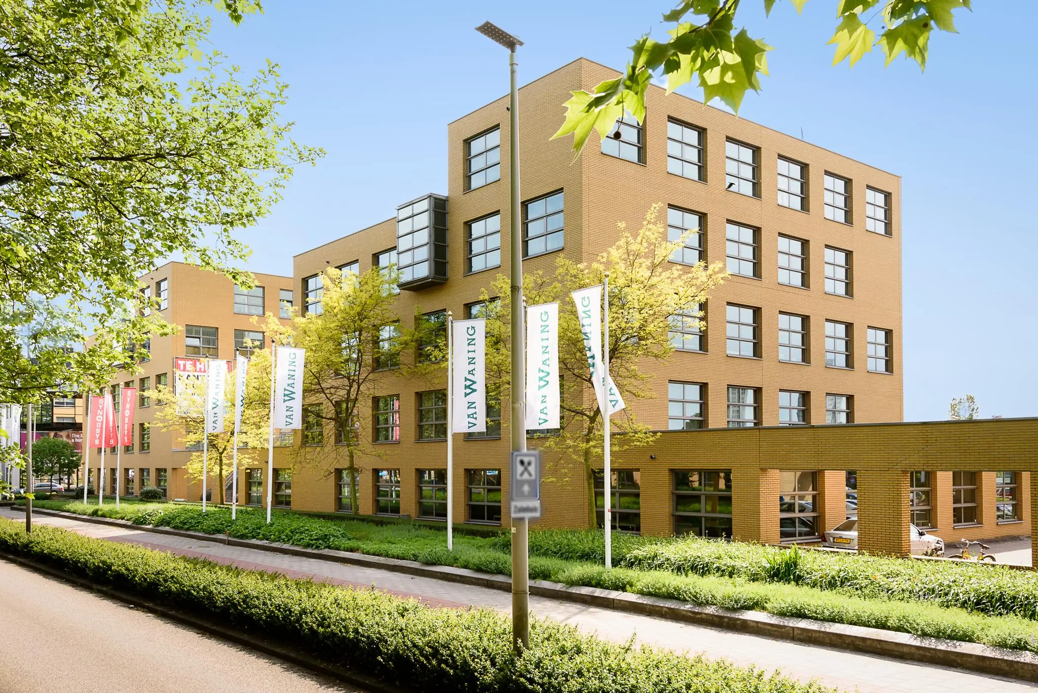 Kantoorgebouw aan de Rivium 2e Straat met vlaggen van Van Waning, omringd door groen en een heldere blauwe lucht.